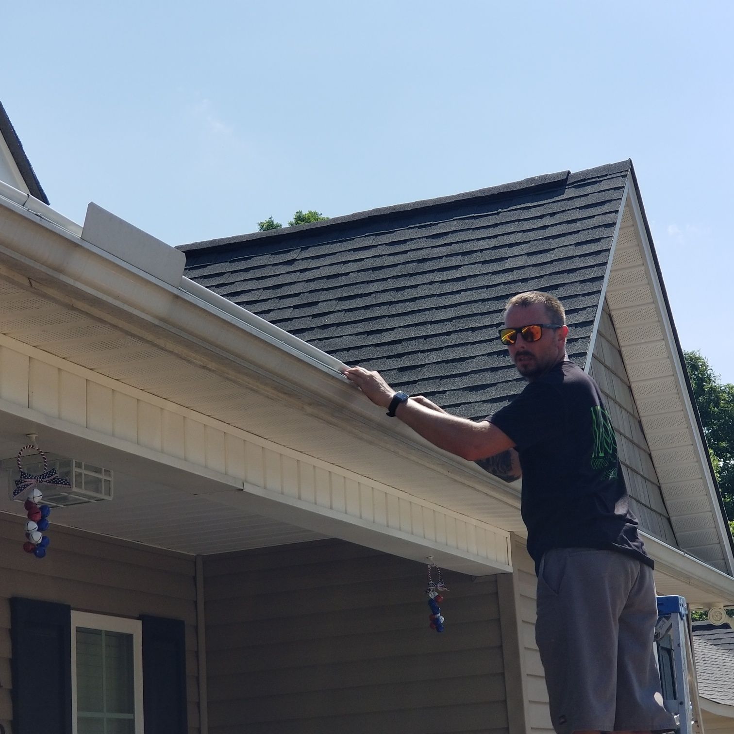 Man on a ladder cleaning gutters on a house under a sunny sky.