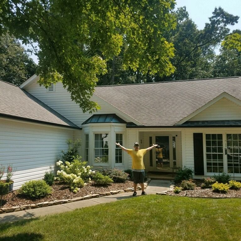 Man in yellow shirt standing in front of white house with arms outstretched; sunny day.