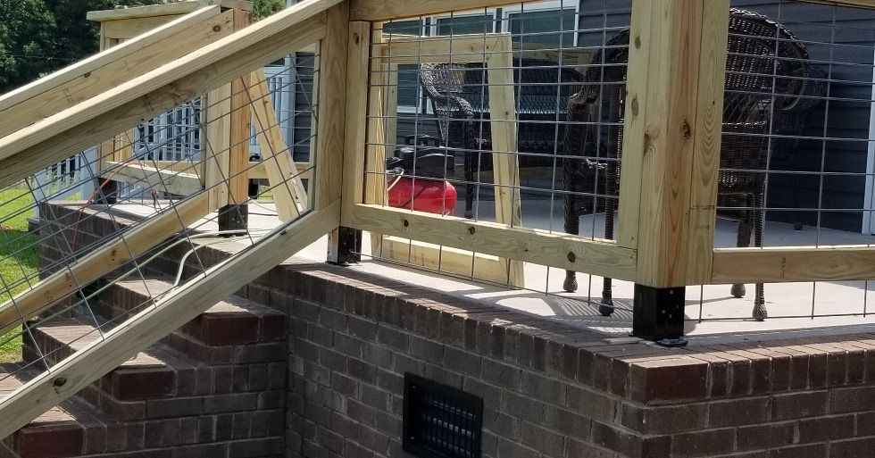 Wooden deck railing with wire mesh, on a brick wall, with a glimpse of outdoor furniture.