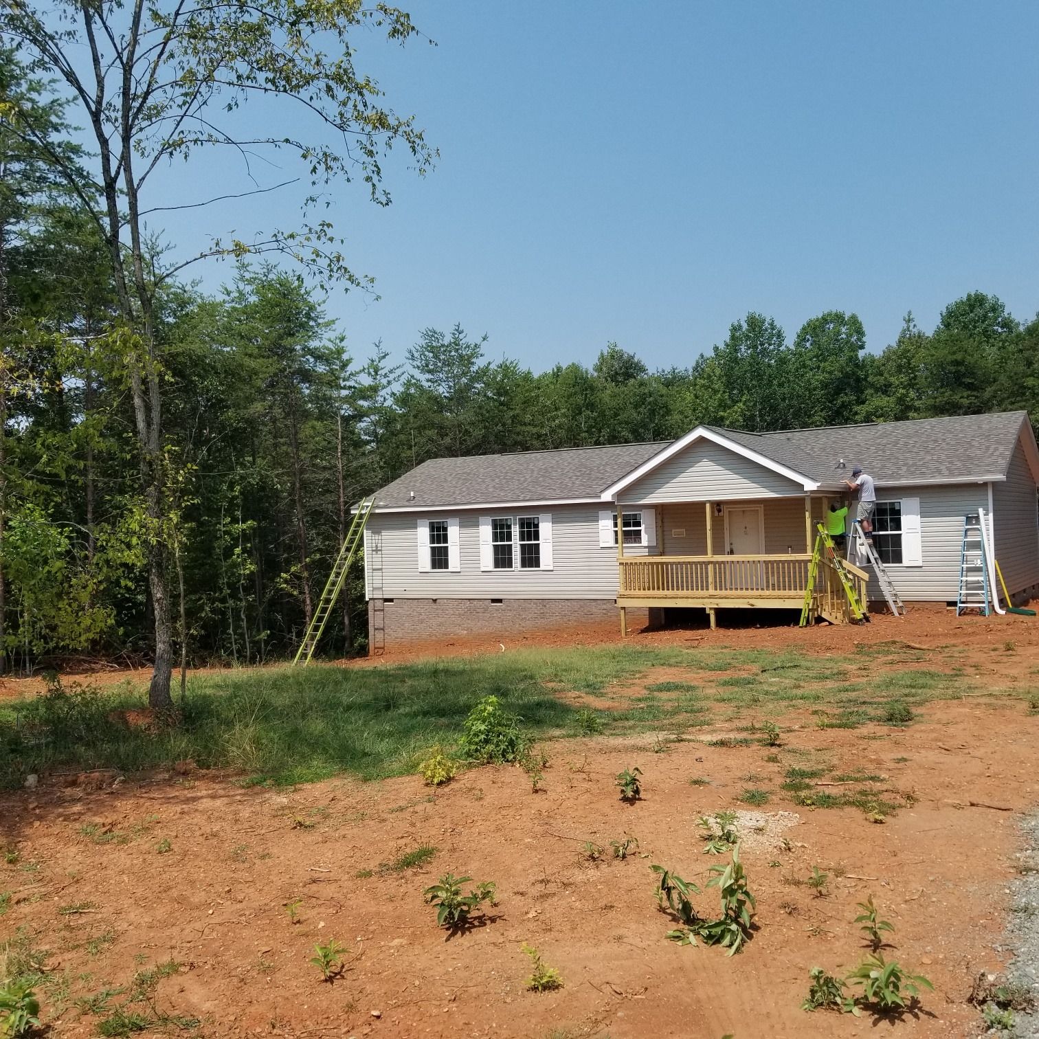 New house under construction, tan siding, small porch, ladders, trees in background, sunny day.
