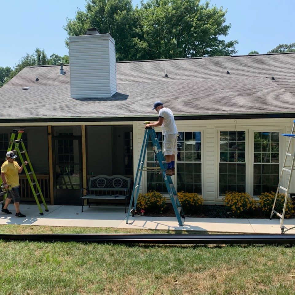 Three people on ladders painting the exterior trim of a house with a gray roof, and white siding on a sunny day.