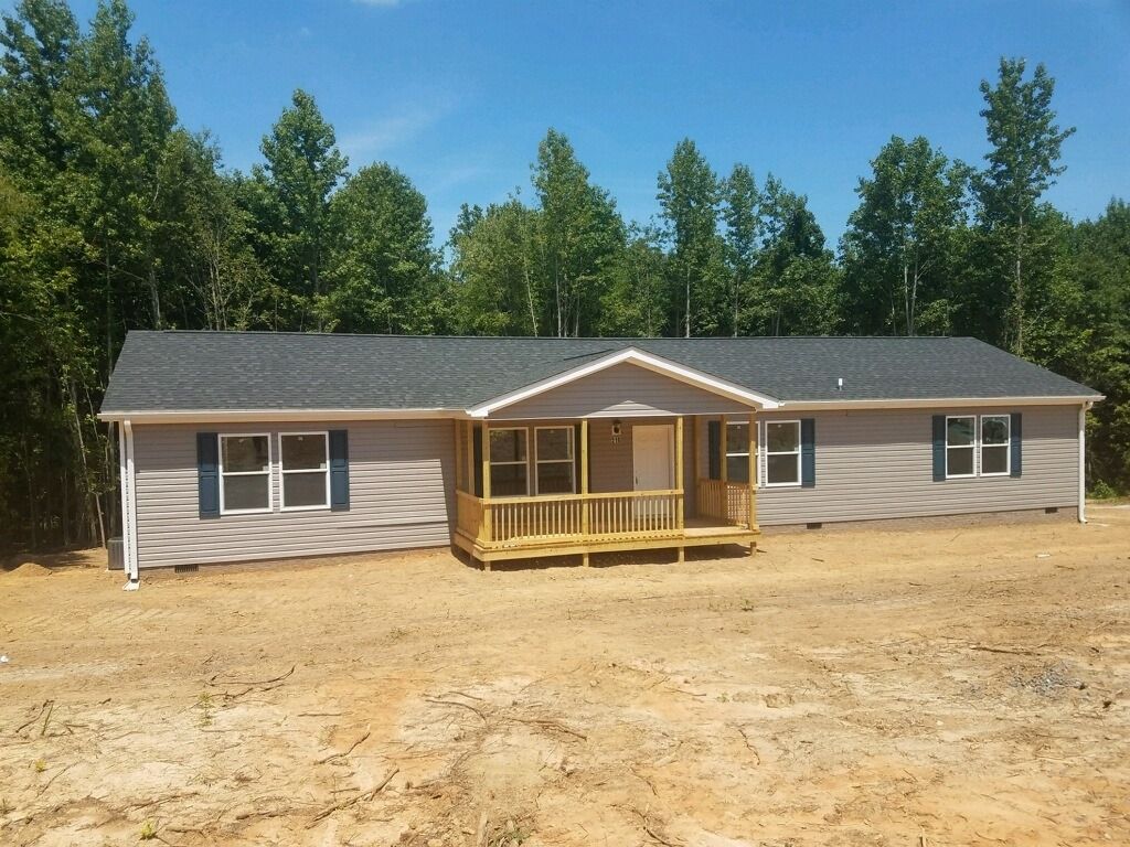 A light gray ranch-style house with a wooden porch and blue shutters in a wooded area under a blue sky.