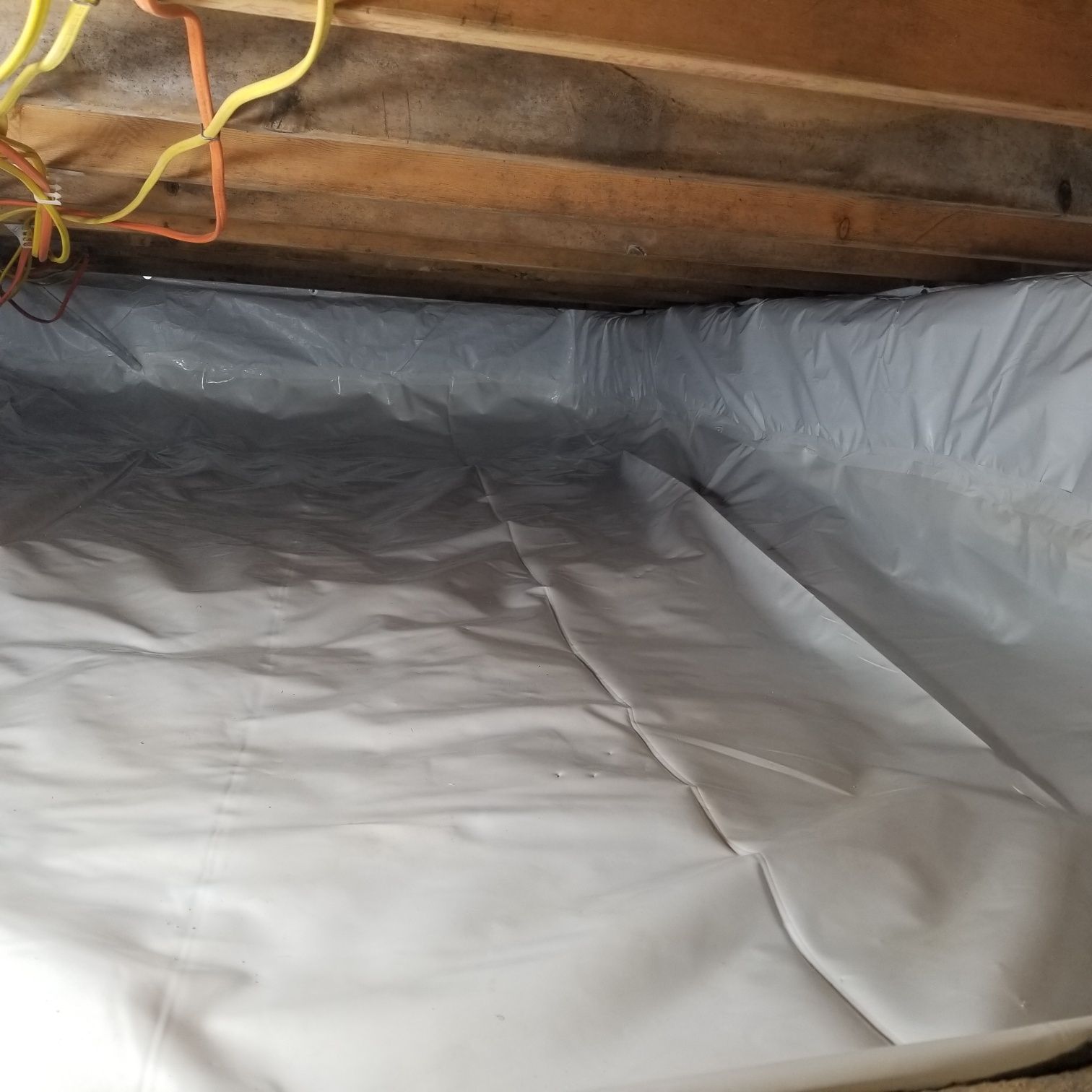 Crawl space with white plastic vapor barrier installed on the floor and walls, wooden beams overhead.