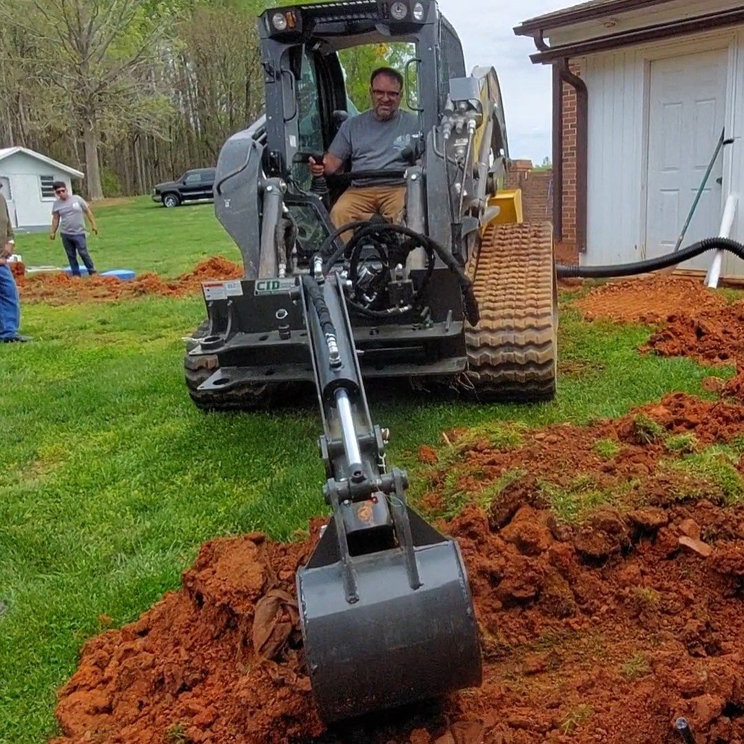 Man operating a small excavator, digging a trench in a grassy yard.