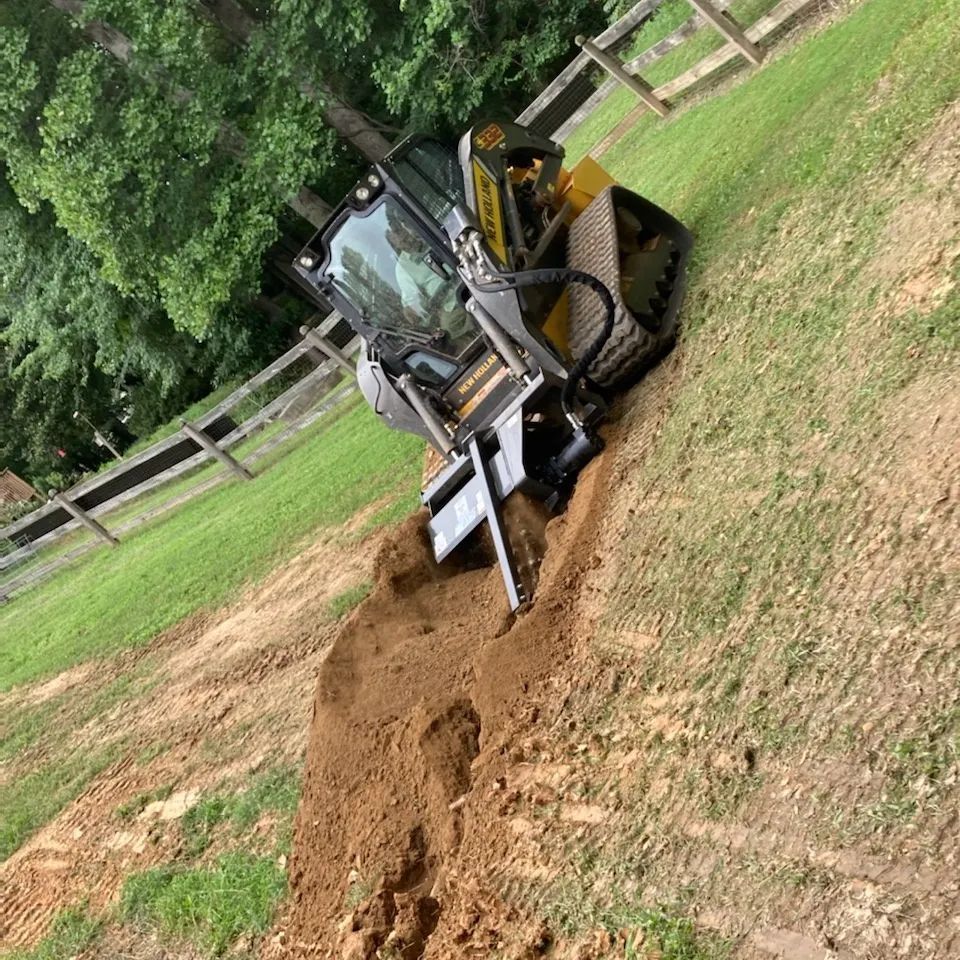 Skid steer tractor digging trench on a grassy slope, near a wooden fence and trees.