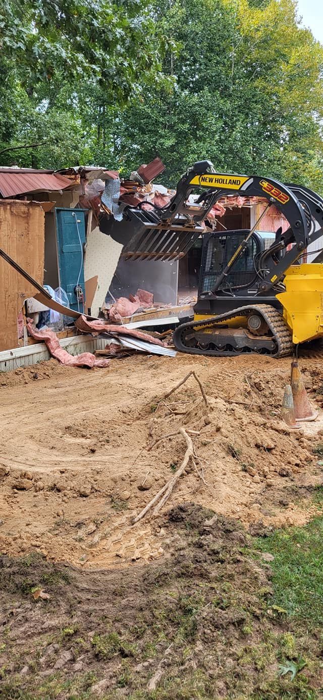 Demolition of a small building by a tracked excavator. Debris and dirt fill the foreground, green trees in the background.