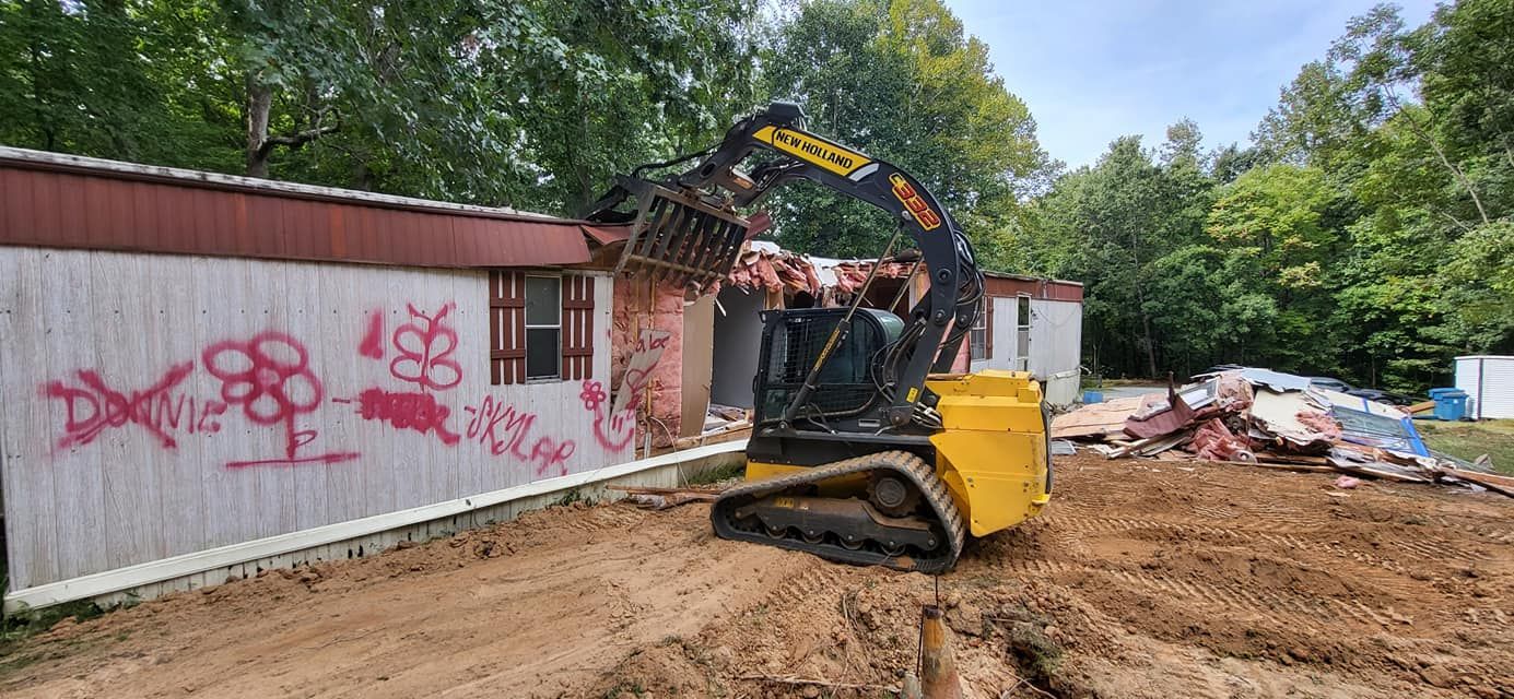 A yellow excavator demolishes a graffiti-covered mobile home. Dirt and debris are in the foreground. Trees in the background.