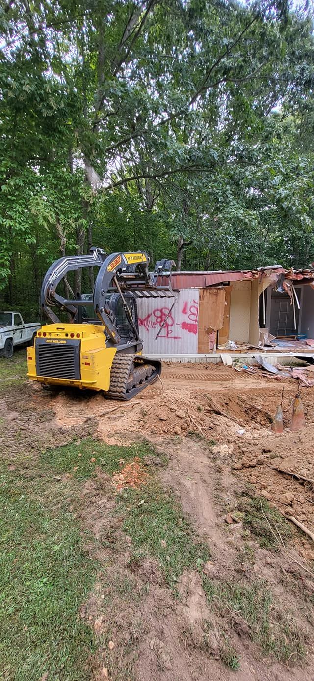 A yellow skid steer demolishes a building, with debris and scattered around. Overcast sky and green trees in the background.