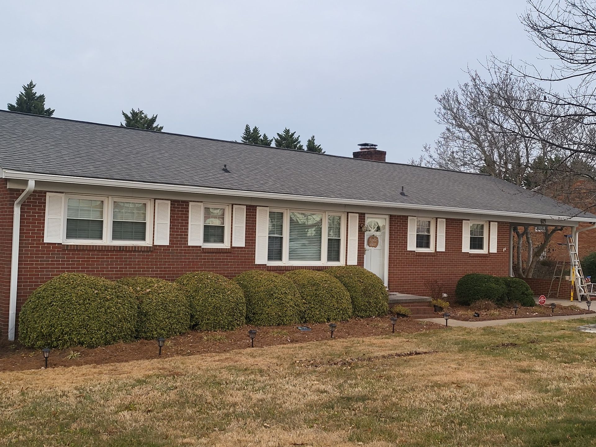 Brick ranch house with white shutters, green bushes, and a gray roof under an overcast sky.