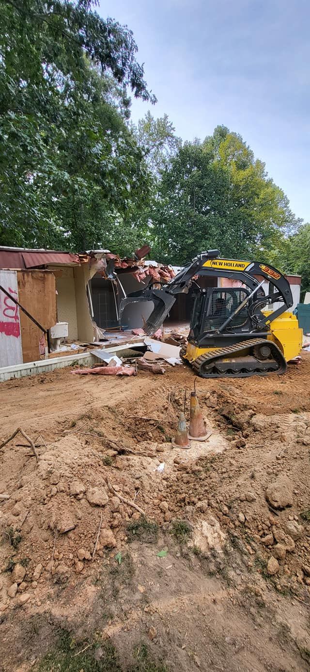 A yellow and black skid steer demolishes a building, surrounded by dirt and trees on a sunny day.