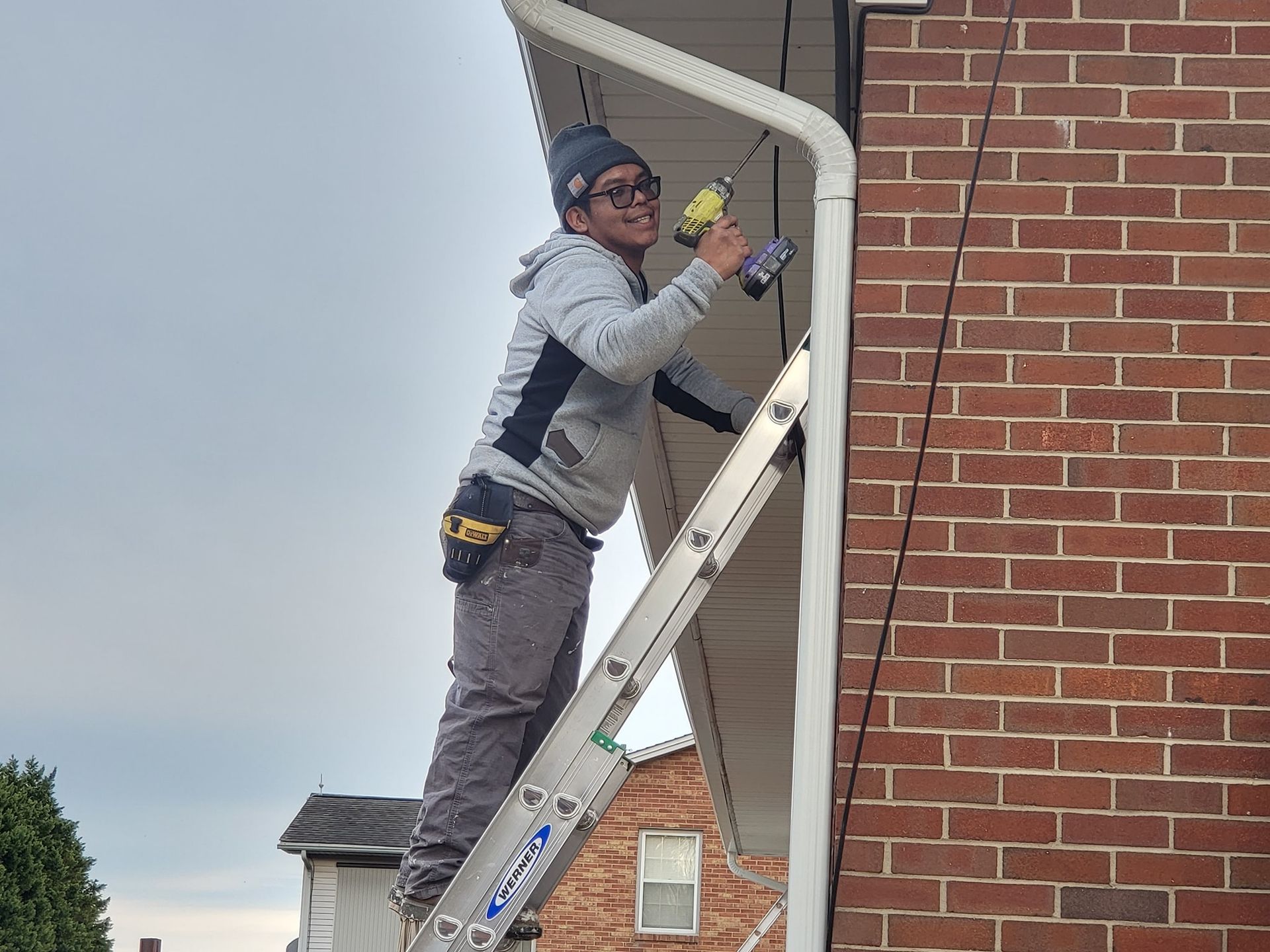 Person on ladder installing gutter with a drill on brick building.