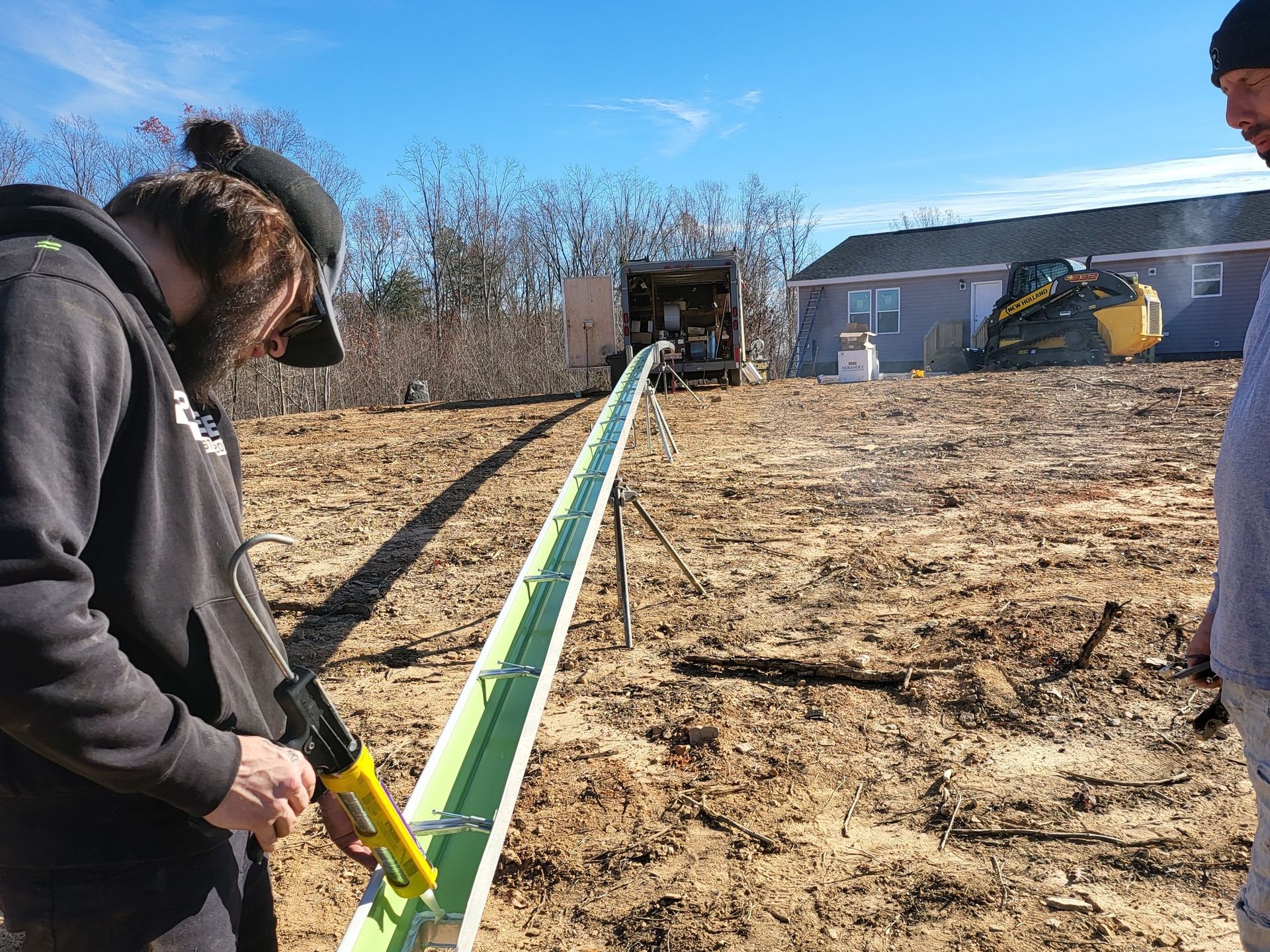 Two men apply sealant to a long, green trough; a trailer is in the background on a brown dirt hill, sunny day.