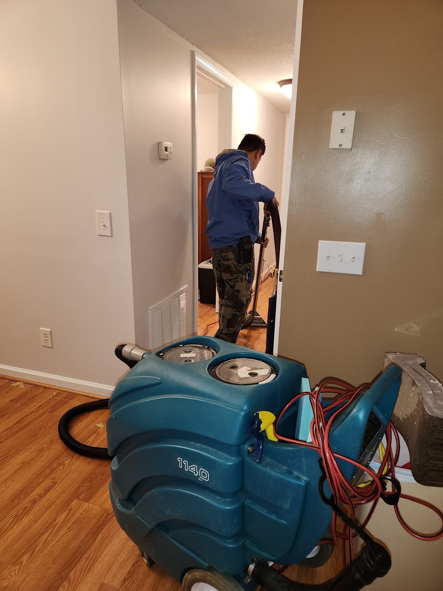 Person carpet cleaning with a machine. The room has light wood floors and tan walls.