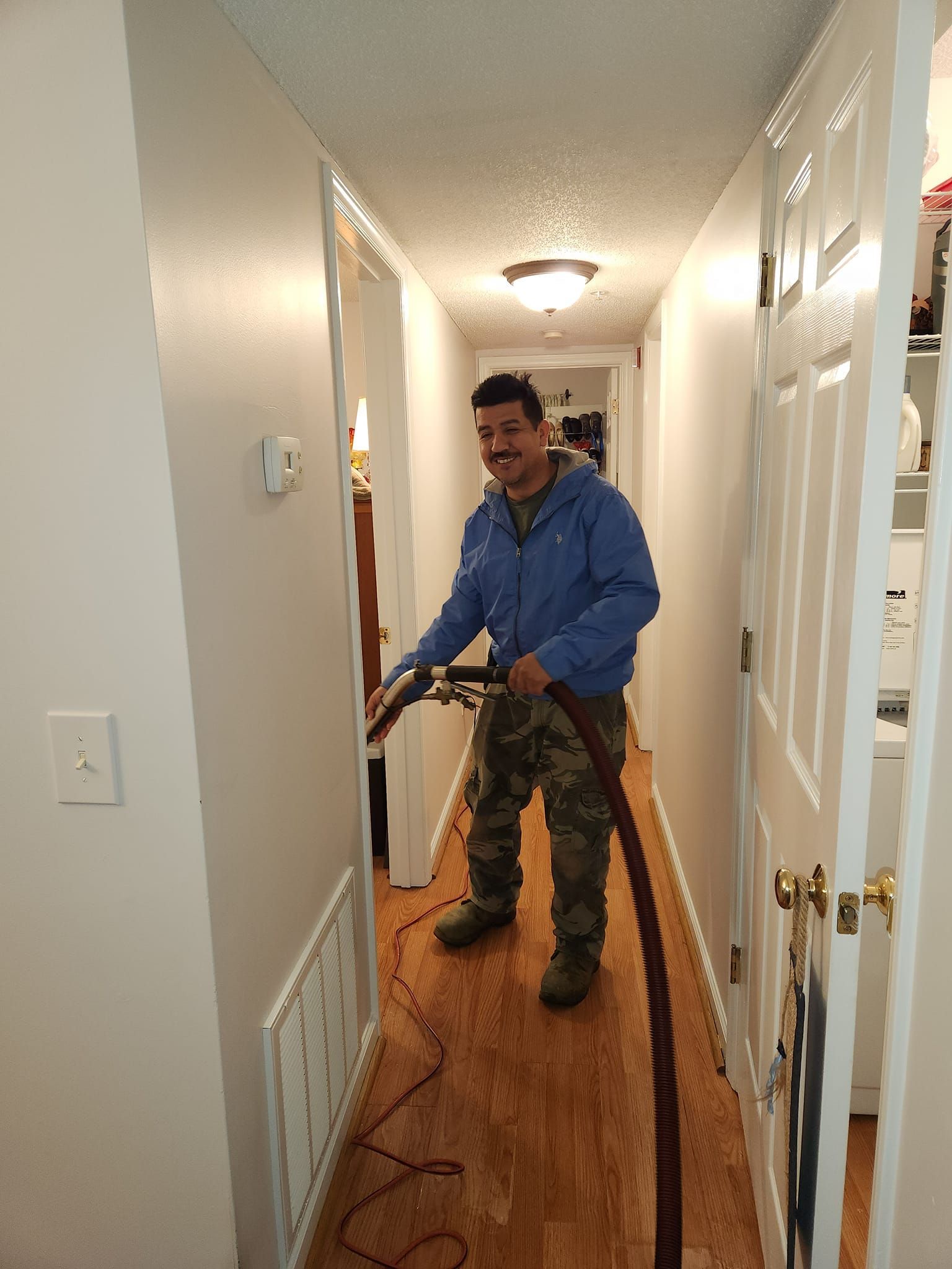 Man vacuuming a hallway with wood-look flooring. He's wearing a blue jacket and camouflage pants.