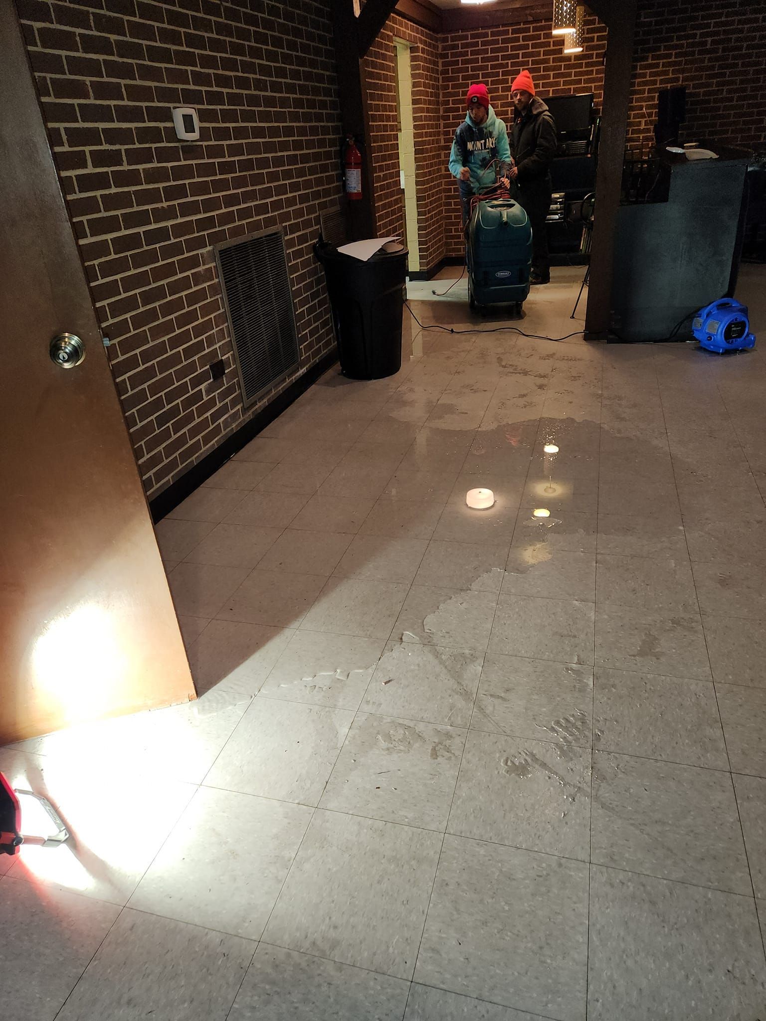 Room interior with two people near a trolley, dark brick wall, trash can, and light on the floor.