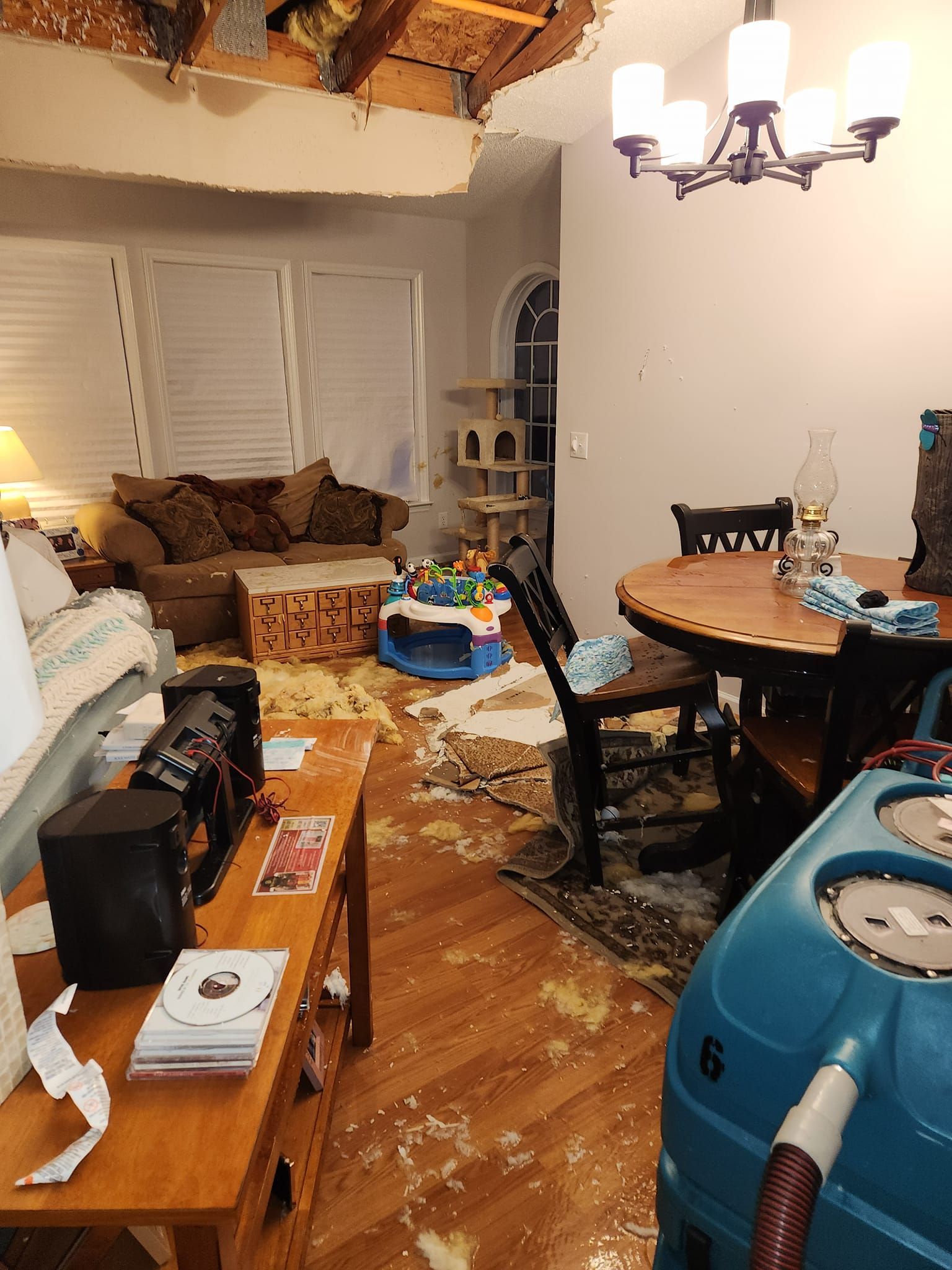 Living room damaged by ceiling collapse; debris on floor and furniture, drying machine visible.