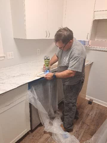 A person applies adhesive to a countertop in a kitchen. They wear a gray t-shirt and work pants.