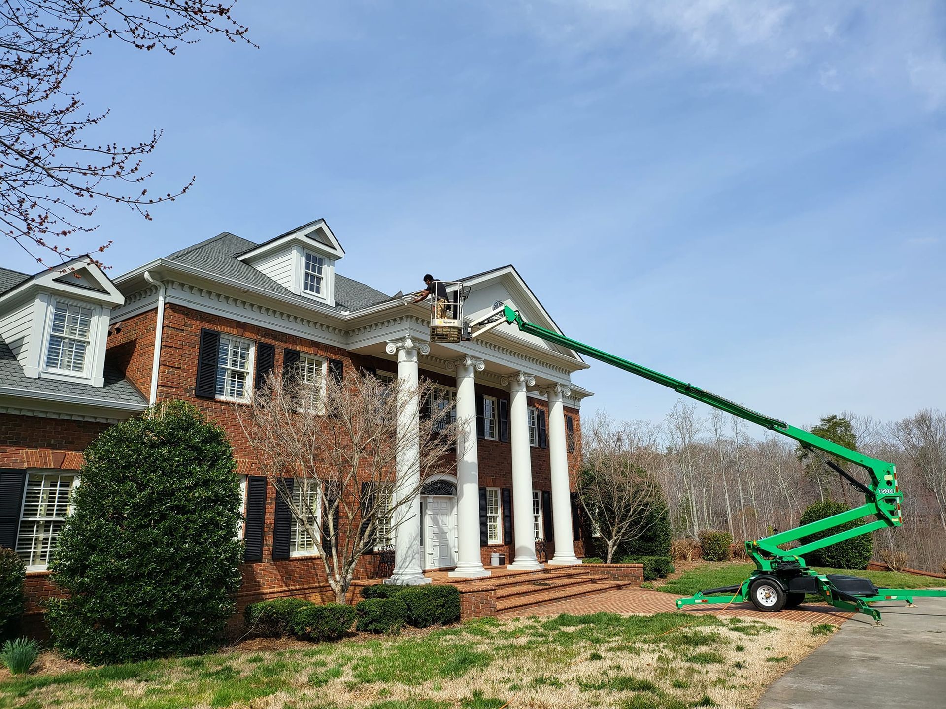 A worker in a green lift is repairing the trim of a two-story brick house with white columns.