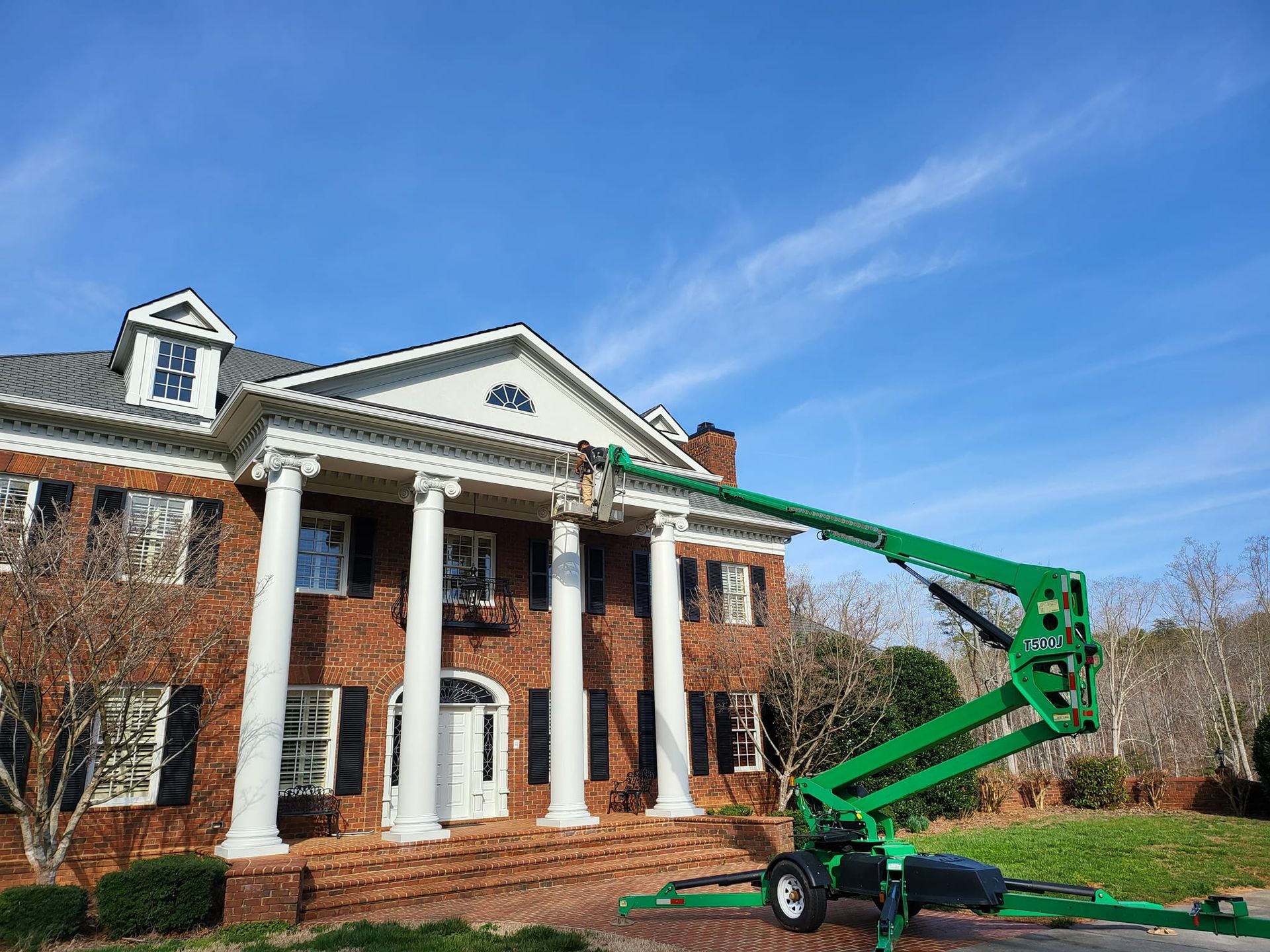 Green lift truck near a brick house with white columns; a worker is at the roofline against a blue sky.