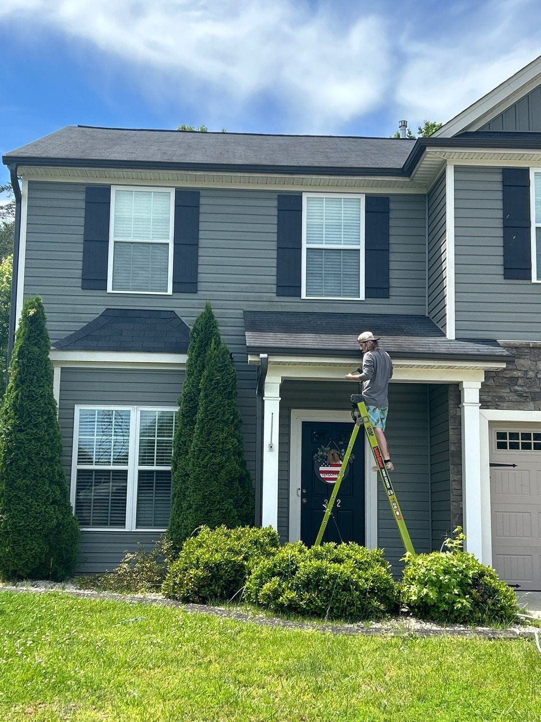 A person on a ladder working on the front of a two-story gray house with black shutters, under a partly cloudy sky.