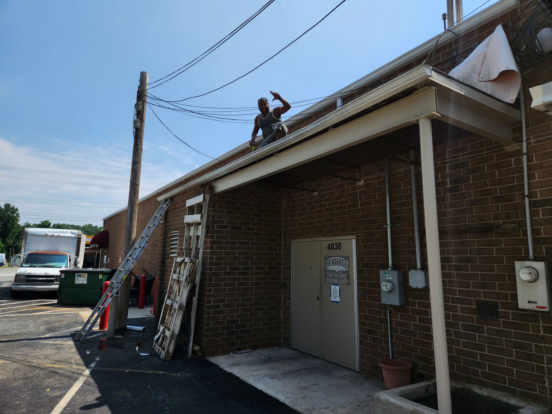 Person on roof near power lines, working on building's brick exterior. Ladder and utility truck nearby on sunny day.