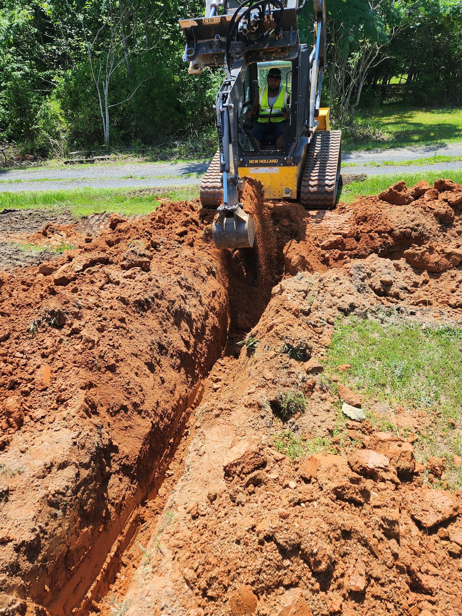 Skid steer digging a trench in reddish-brown soil in a grassy yard. Operator visible in the cab.