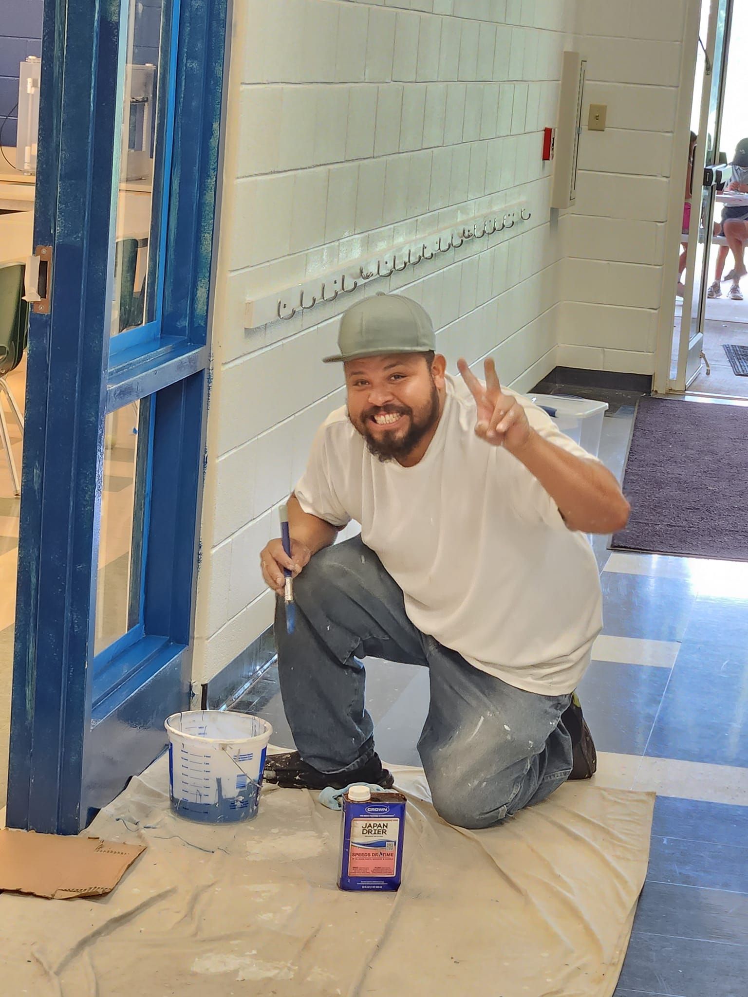 Man painting blue window frame, giving peace sign. White wall, paint bucket, solvent visible.