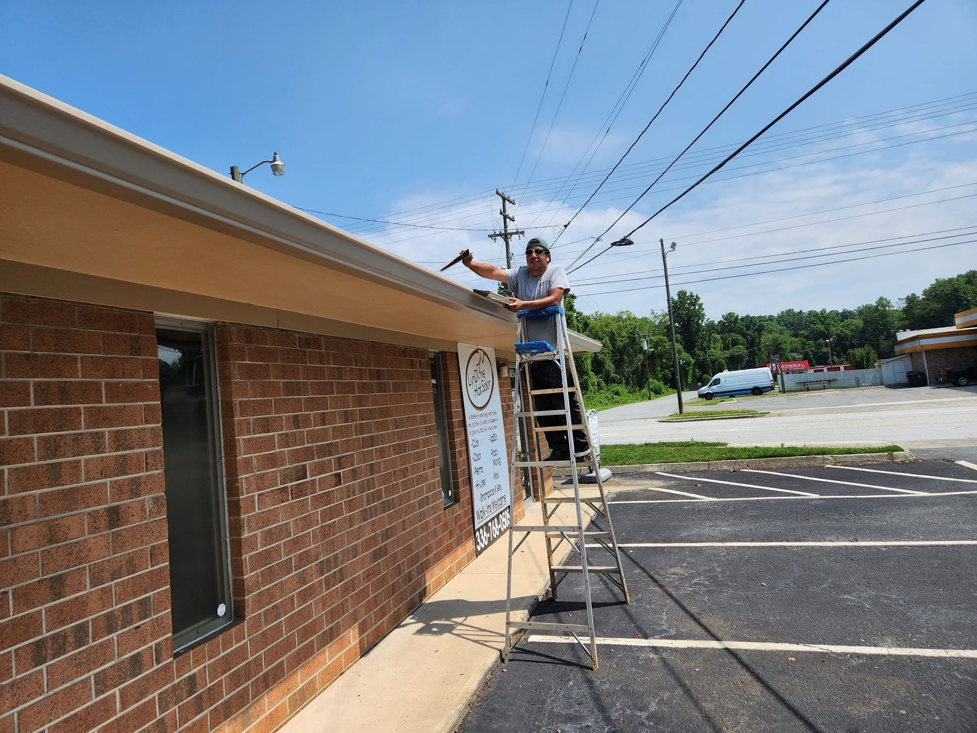 Person on ladder working on the gutter of a brick building under a sunny, blue sky.