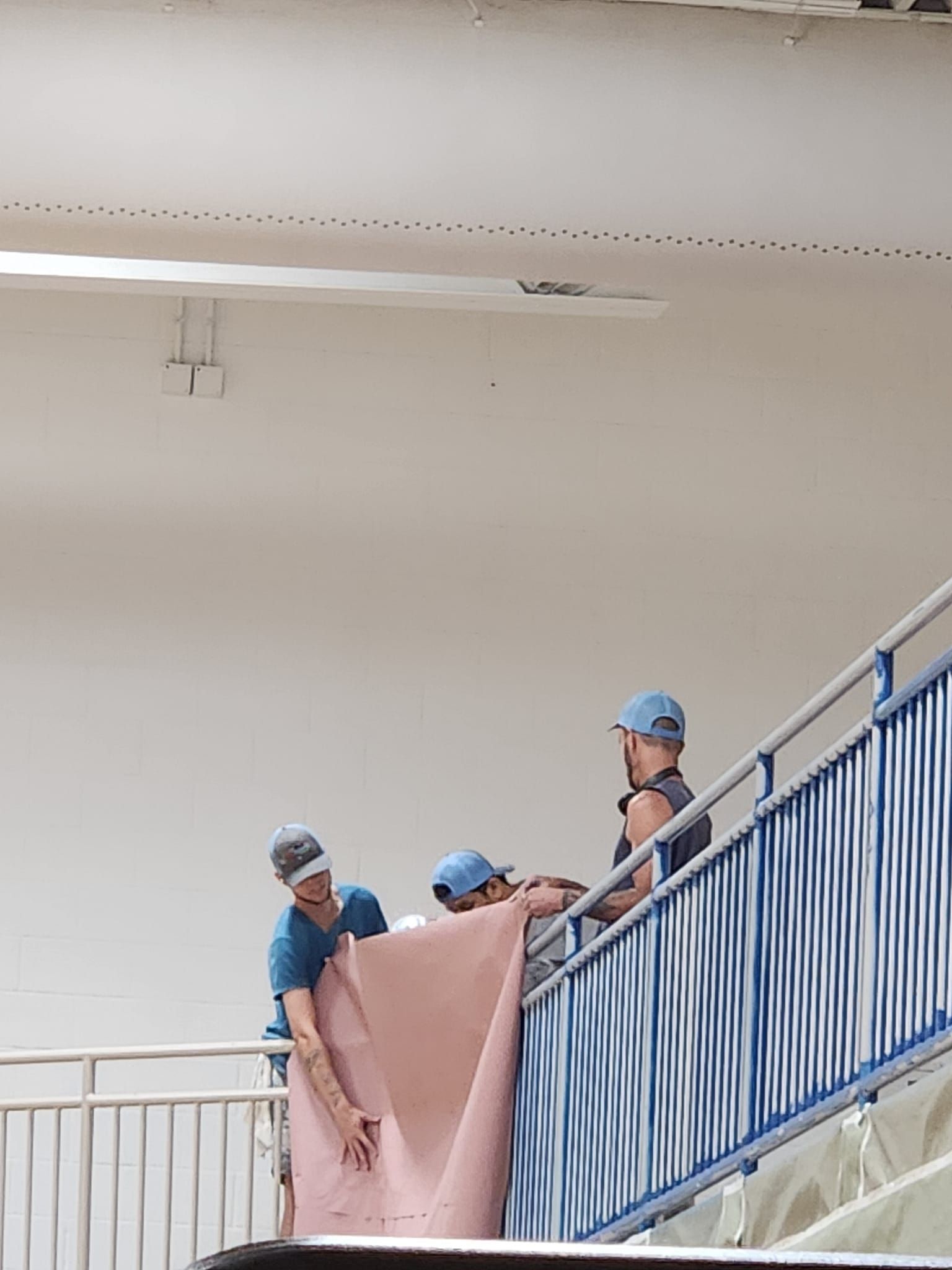 Three people carrying a rolled-up pink rug down a flight of stairs with blue railings.