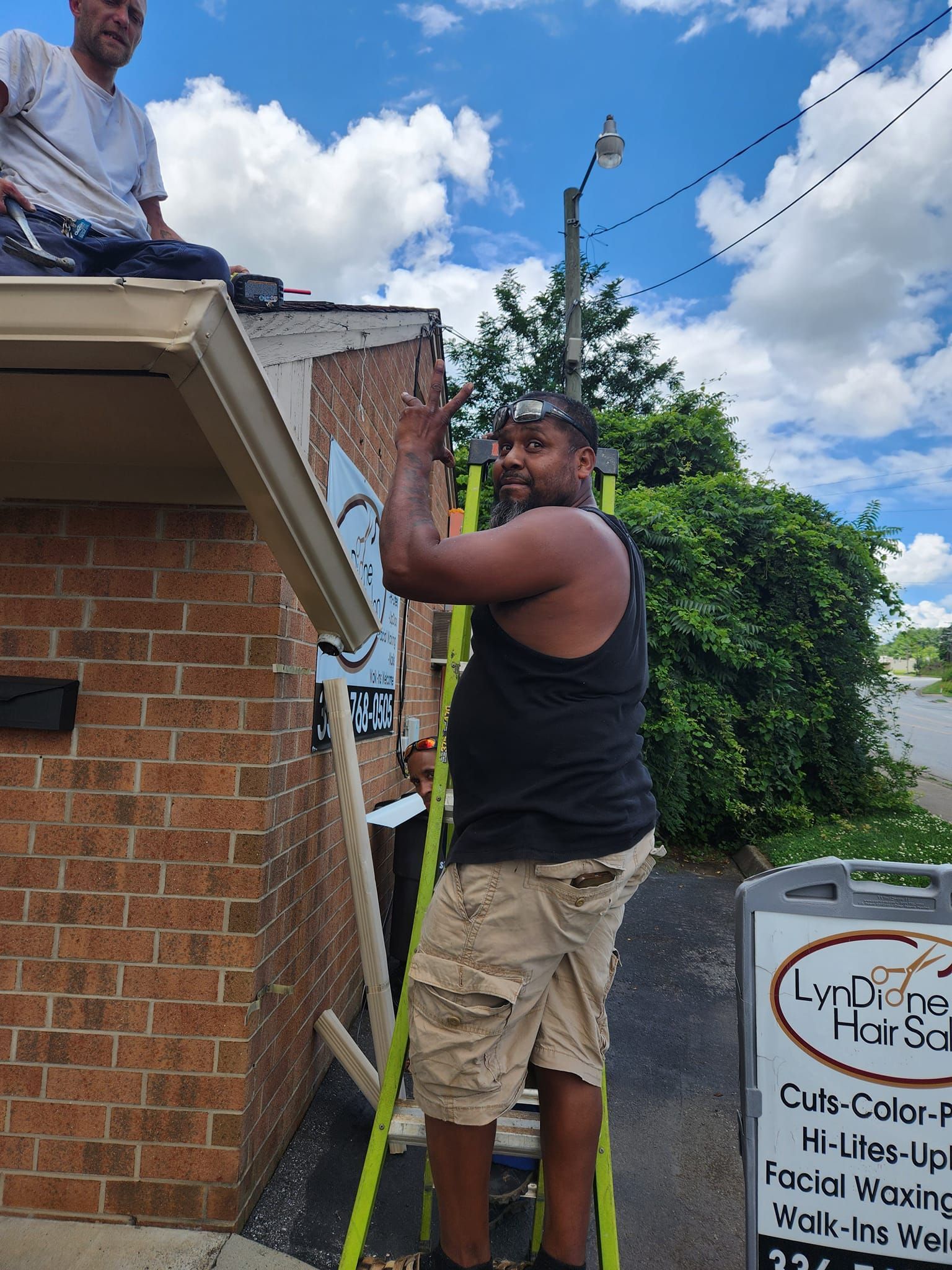 Man on ladder outside a brick building waving, another person on roof, sunny day.