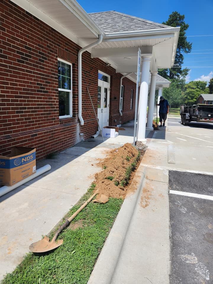 Exterior view of a brick building with a trench dug along the walkway. A person stands construction equipment is present.