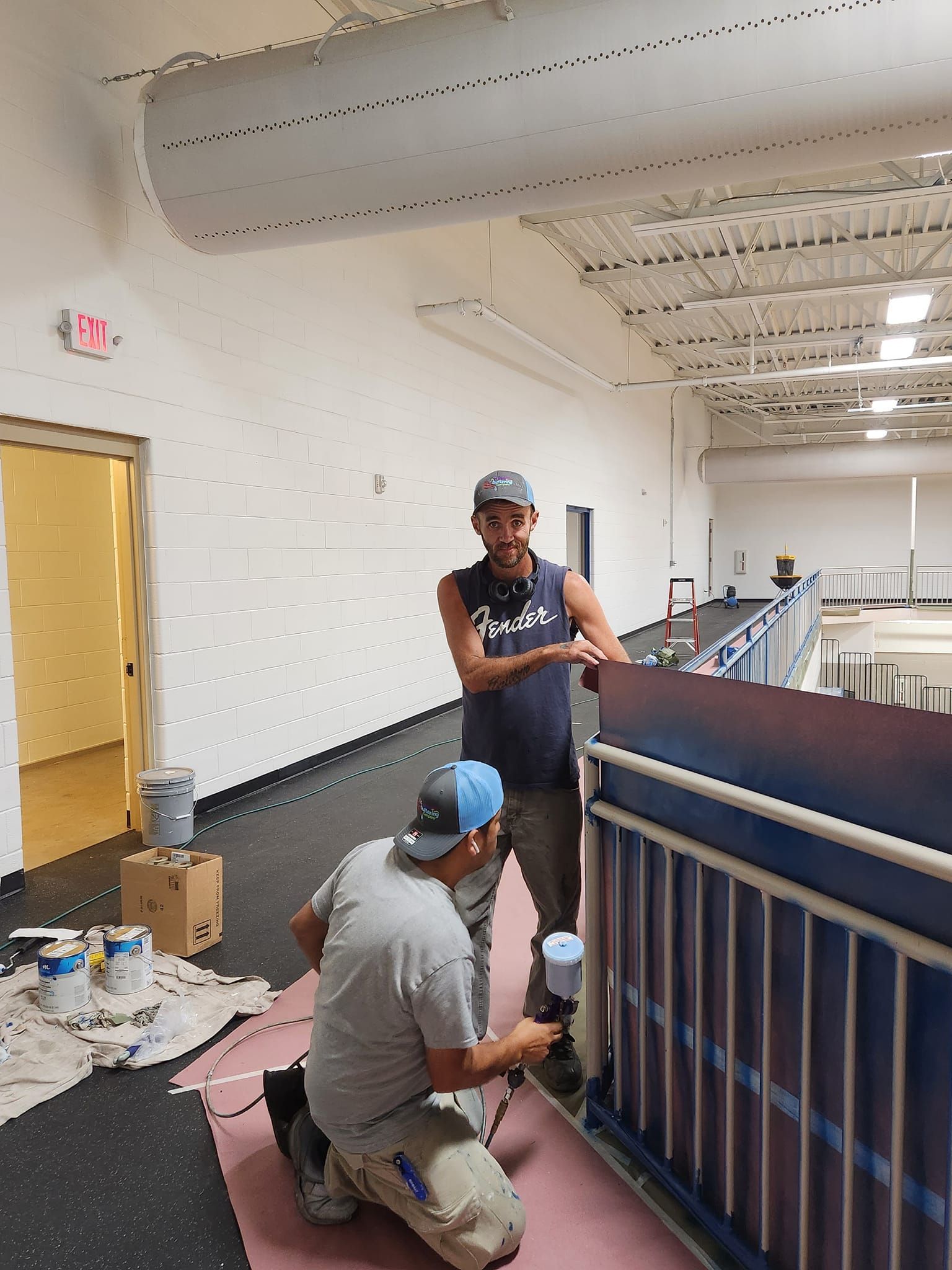 Two people painting an indoor railing. One sprays blue paint, the other watches.