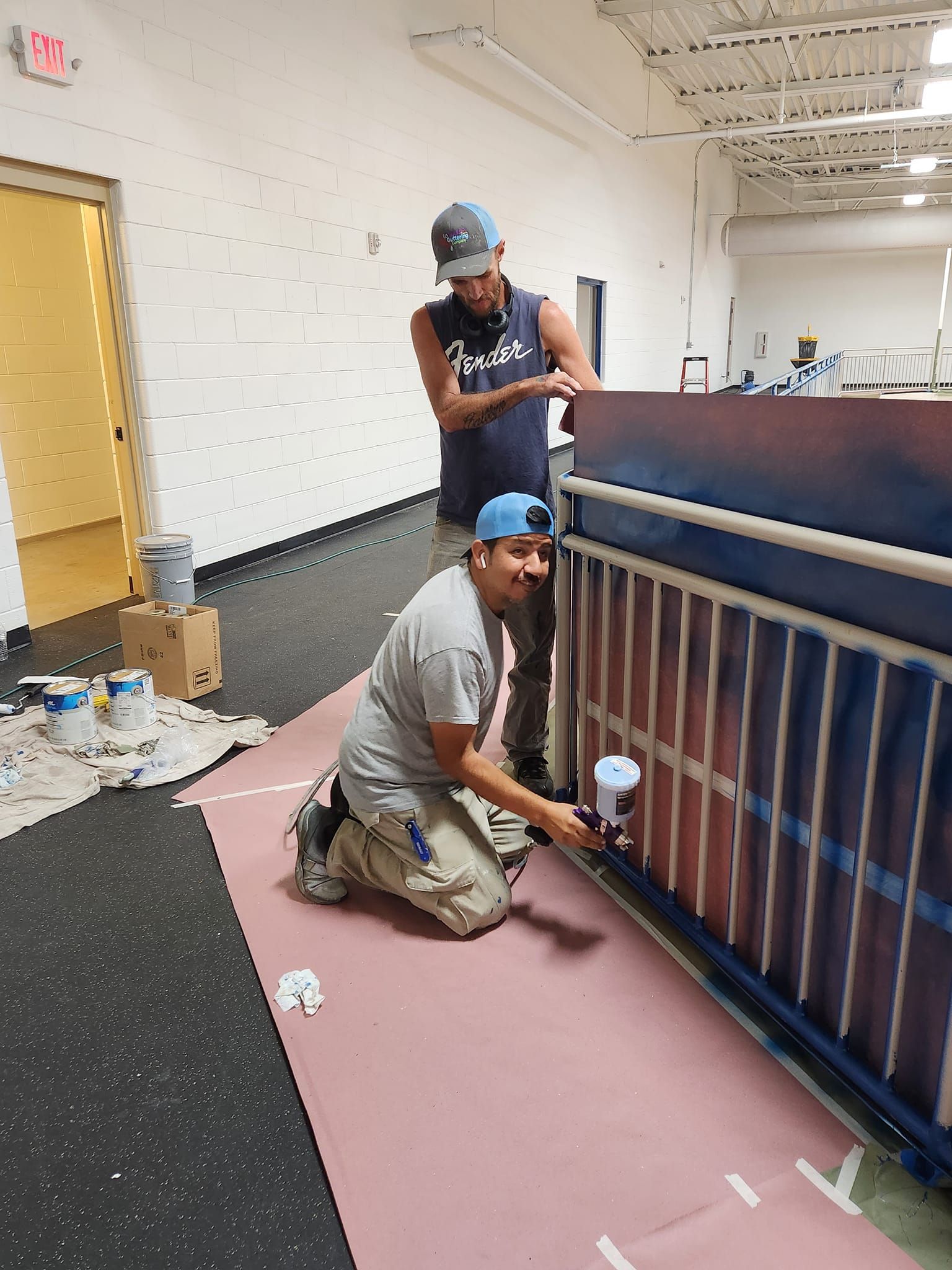 Two people painting a metal railing. One kneels using a spray gun; the other stands nearby.