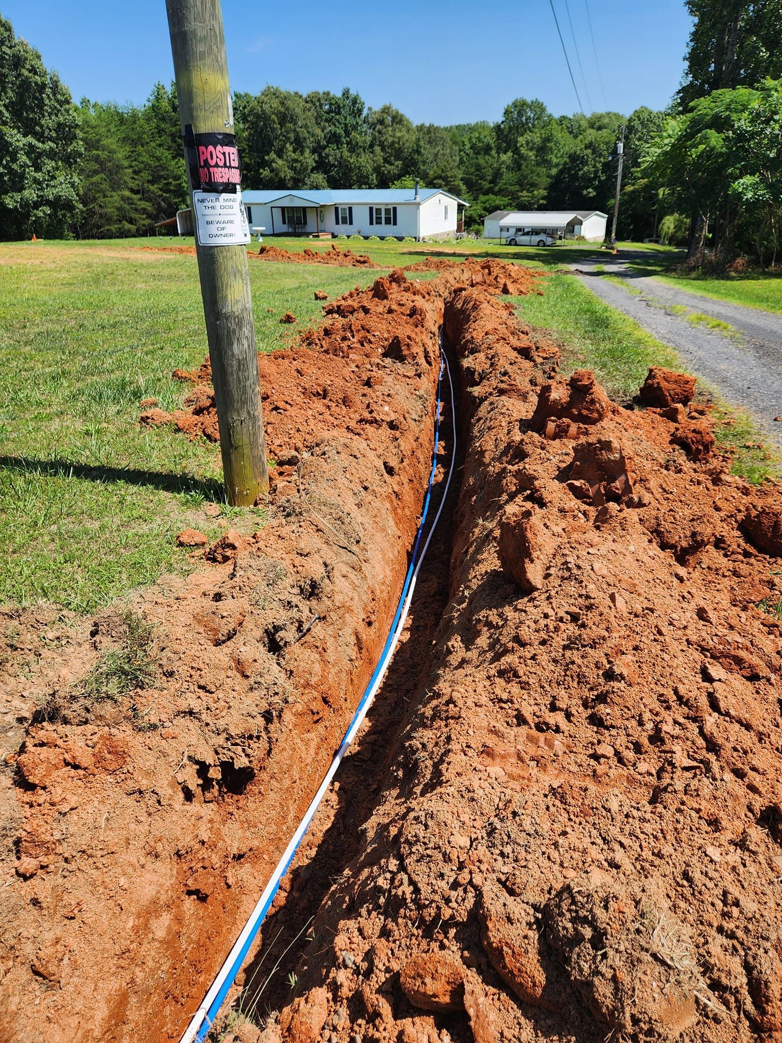 A trench dug along a grassy area next to a dirt road, with a utility pole and houses visible.