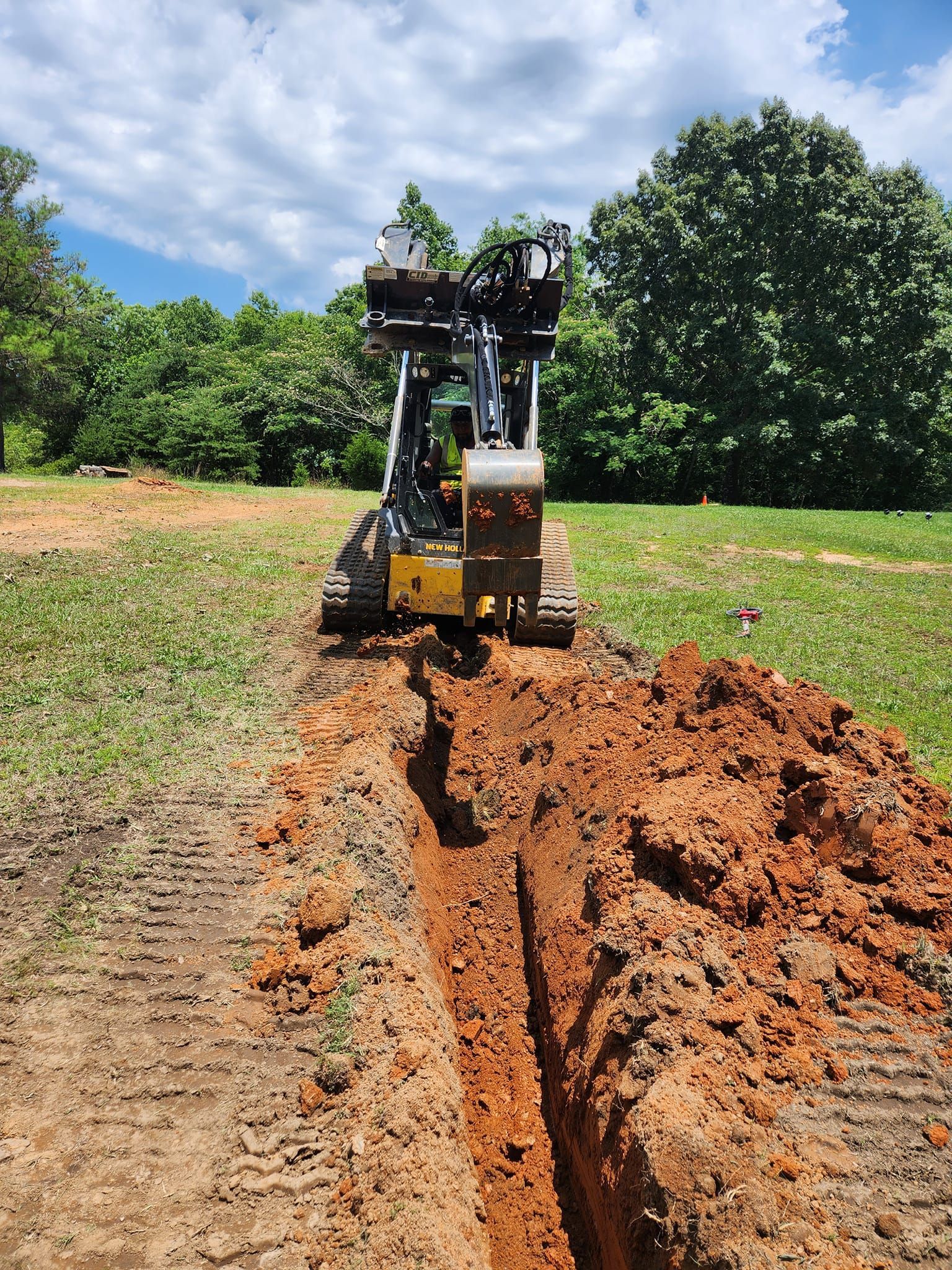 Yellow trencher digging a trench in reddish-brown soil in a grassy field on a sunny day.