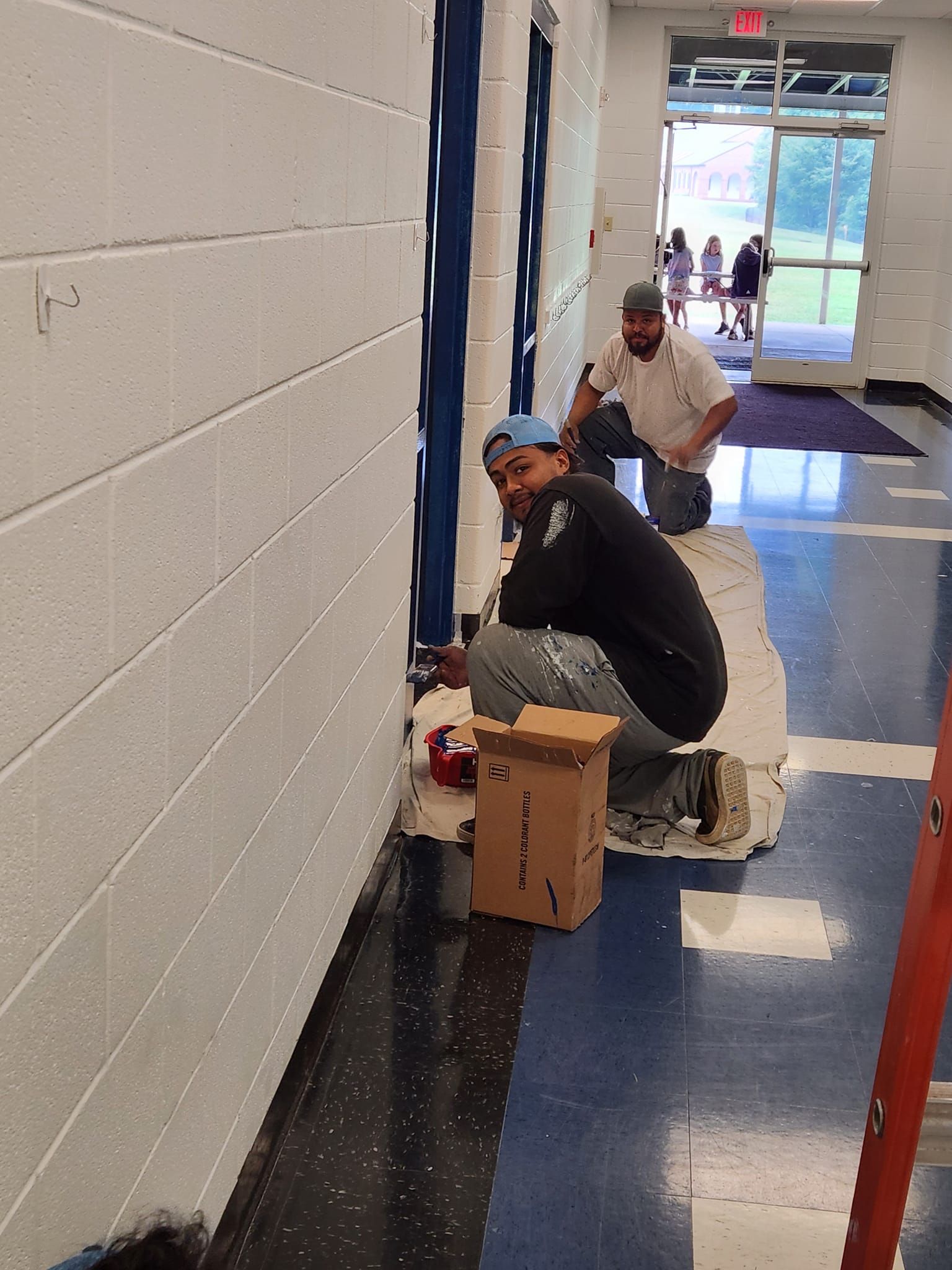 Two men painting blue stripes on a white wall in a hallway. One crouches, the other kneels.