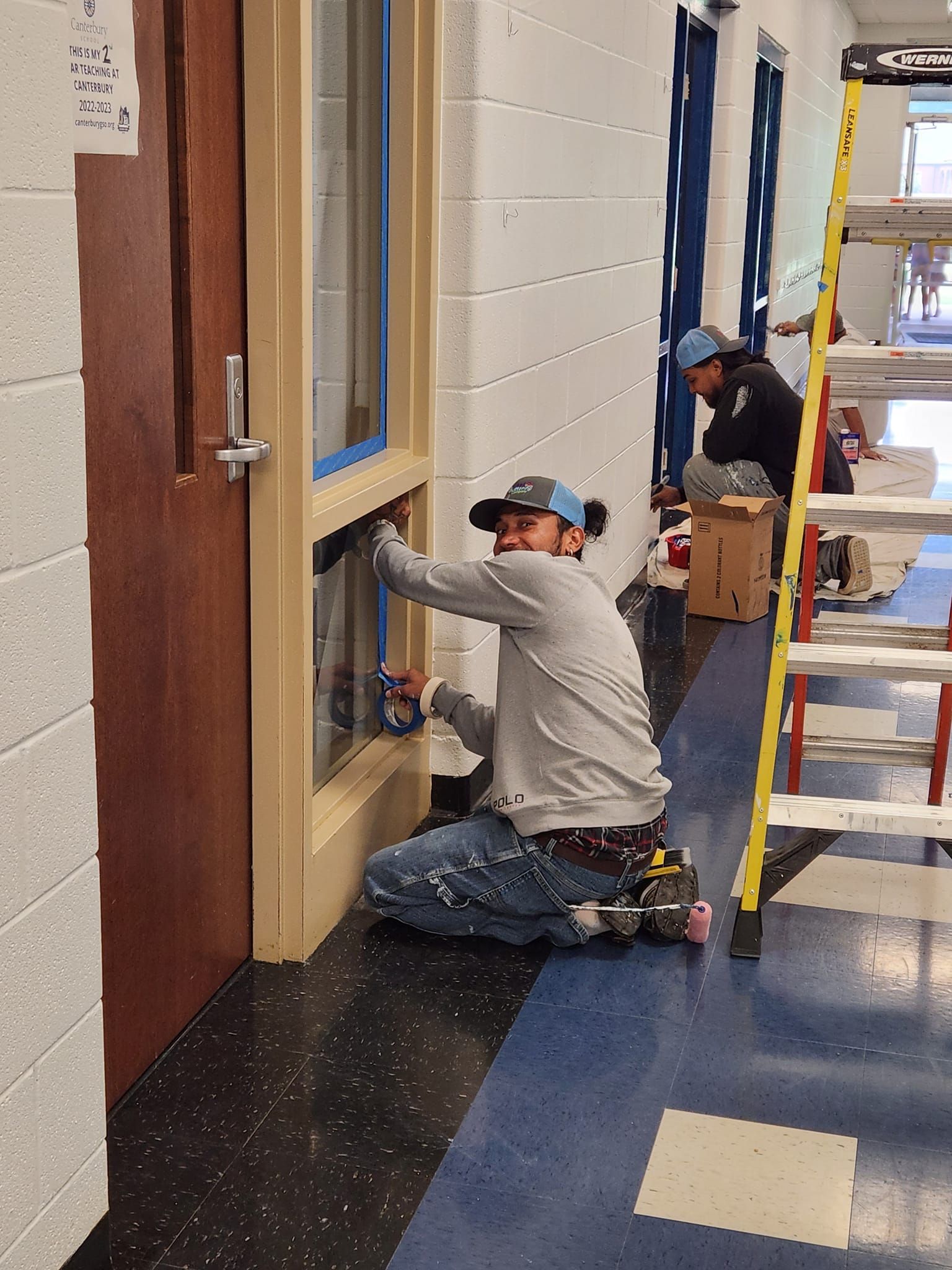 Person kneels, painting window frame in a hallway. Another person works in the background by a ladder.
