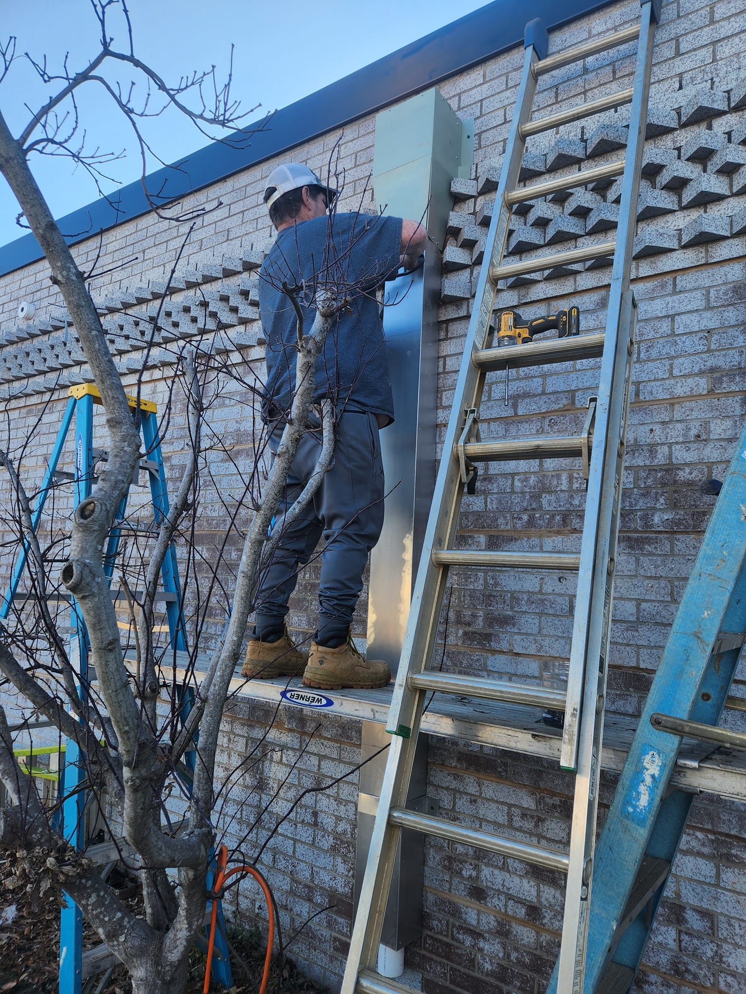 Worker on ladder installing a metal panel on a brick building exterior.