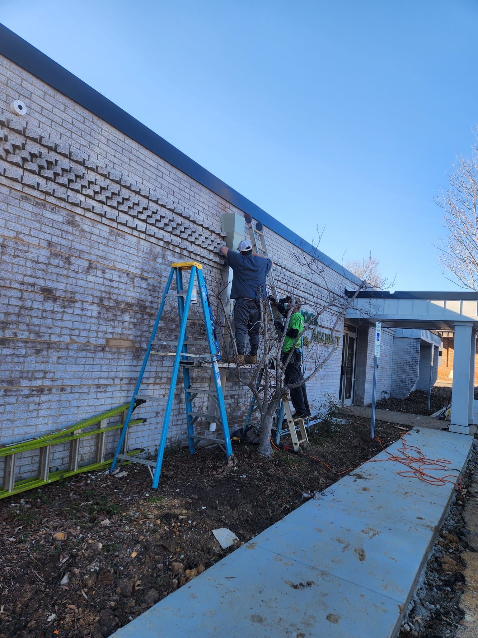 Two workers installing equipment on a brick building, one on a ladder, sunny day.