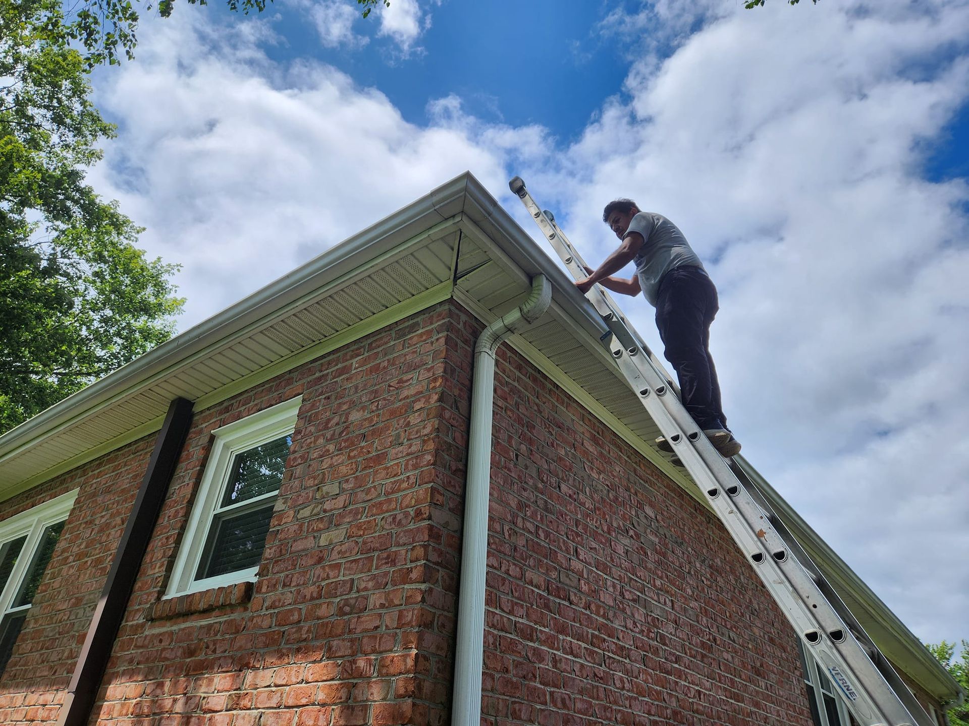 Person on ladder cleaning gutters of a brick house under a cloudy sky.