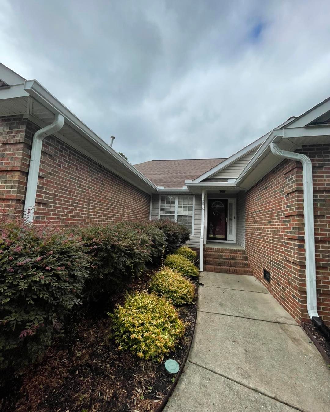 Brick house entrance with walkway, bushes, and cloudy sky.