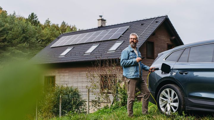 Man charging electric car in front of a house with solar panels on the roof.