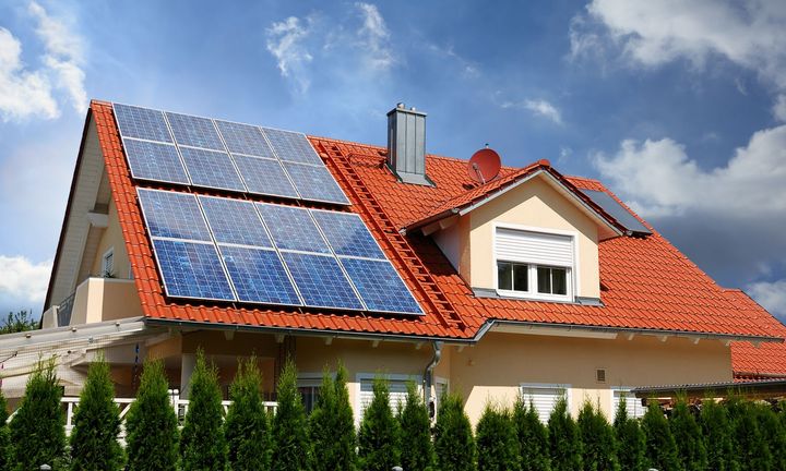 House with solar panels on a red tile roof; blue sky, green bushes in front.