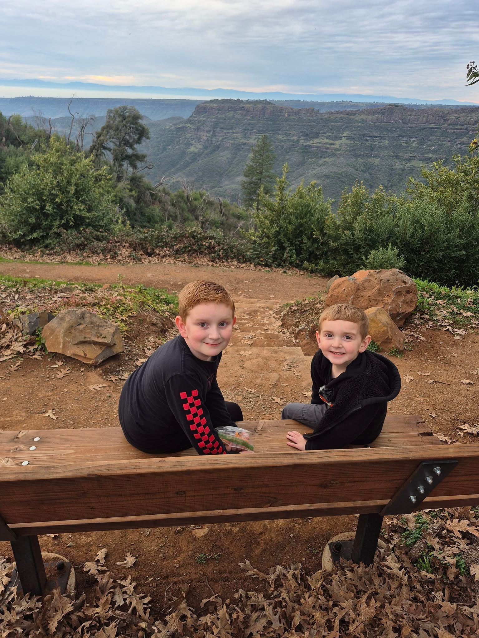 Two children sit on a bench overlooking a canyon under a cloudy sky.