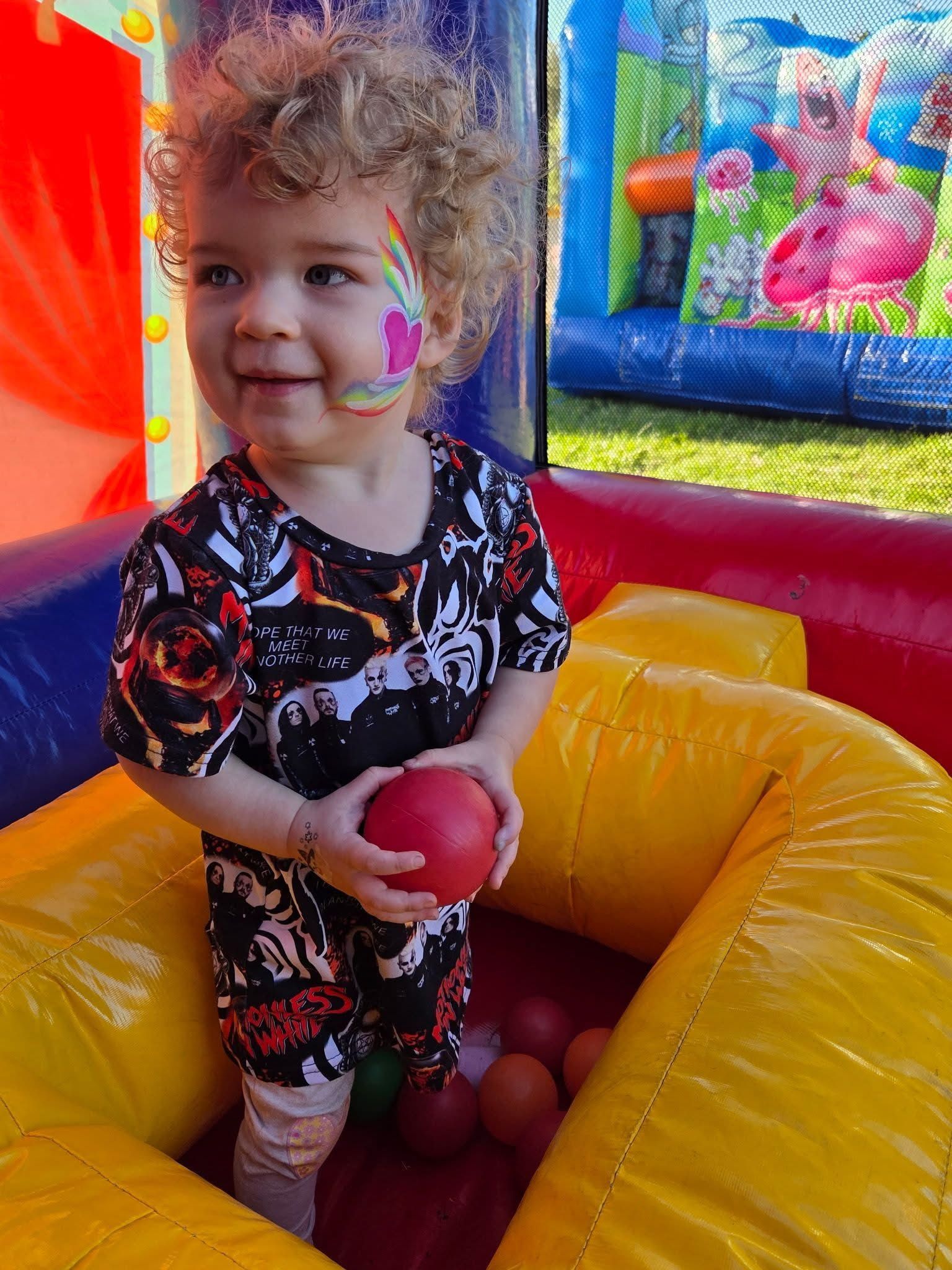 Child in a colorful bounce house holding a red ball, with face paint and a purple inflatable background