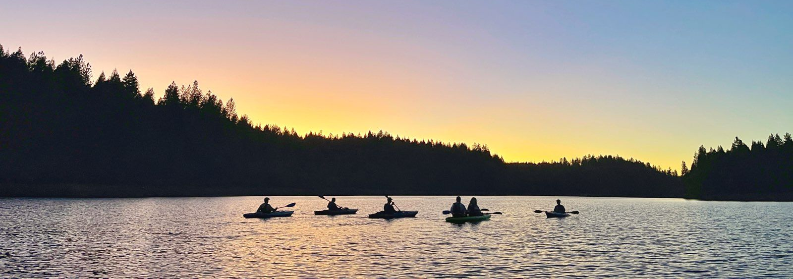 Kayakers on a lake at sunset. Silhouette of people in boats, trees on shore, orange sky.