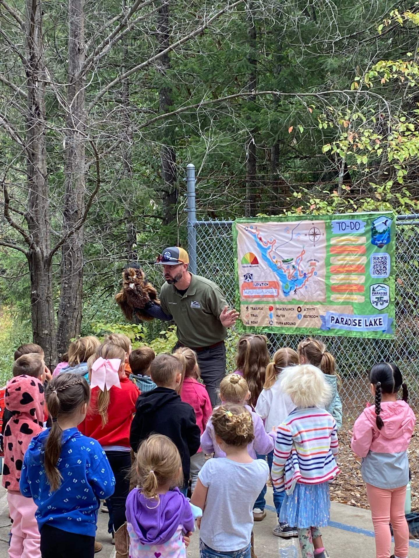 A man with a bird puppet speaks to a group of children near a park map on a fence.