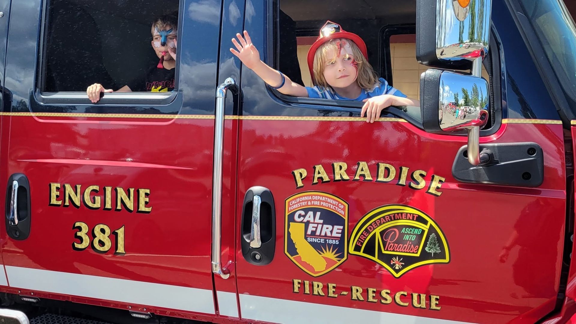 Children waving from a red fire rescue truck labeled “Paradise Fire-Rescue” and “Engine 381”