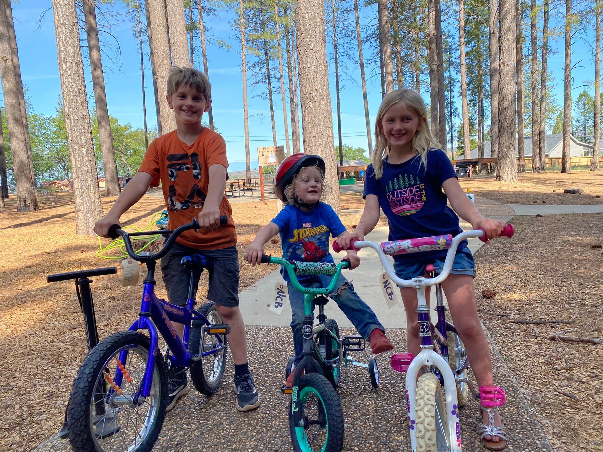 Three children smiling on bikes in a wooded area; blue sky.