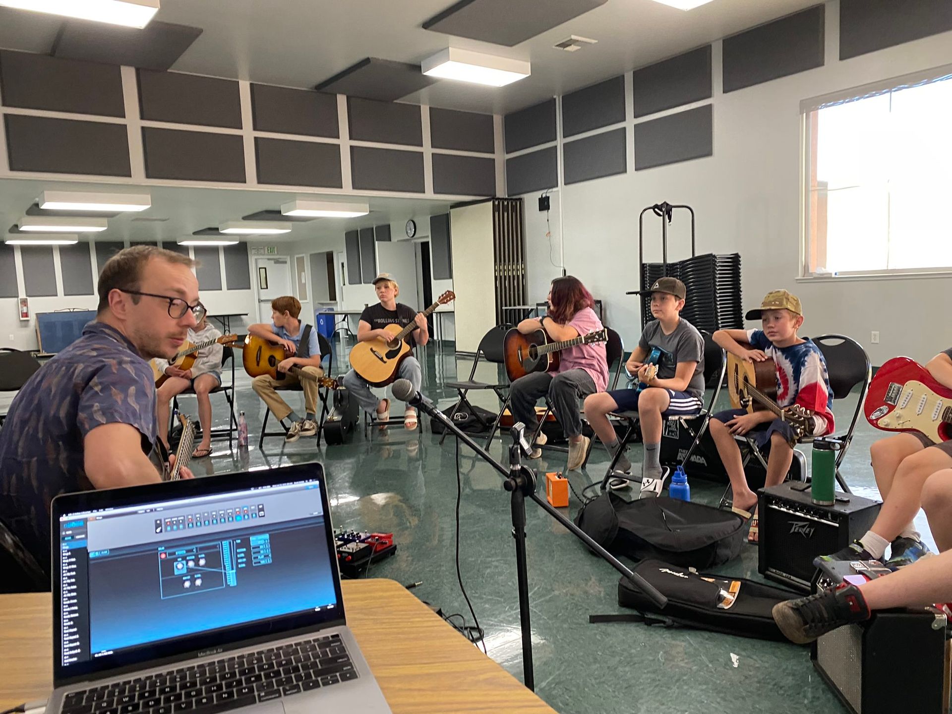 A music class; a teacher and students sit, playing guitars in a room with soundproofing. A laptop is in the foreground.